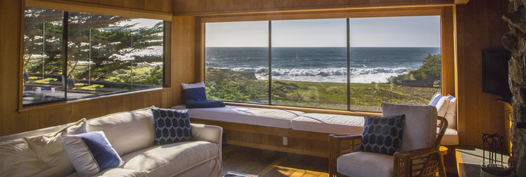 Interior of a room showing a chair, sofa and window seat with view of the ocean through a large window.