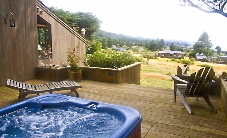 A deck next to a home with hot tub, chair, chiase, and view of the garden beyond the deck.