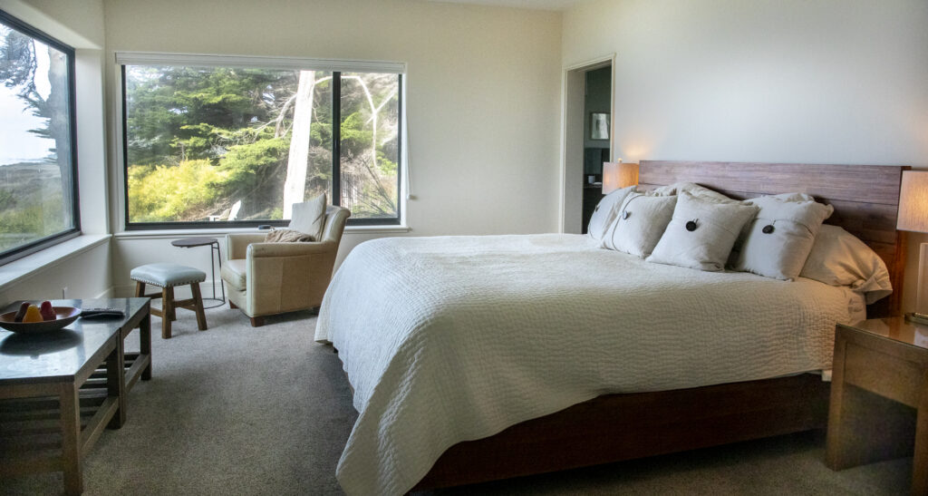 A bedroom with bed, chair, footstool, table, two lamps and a view of the garden out a large window.